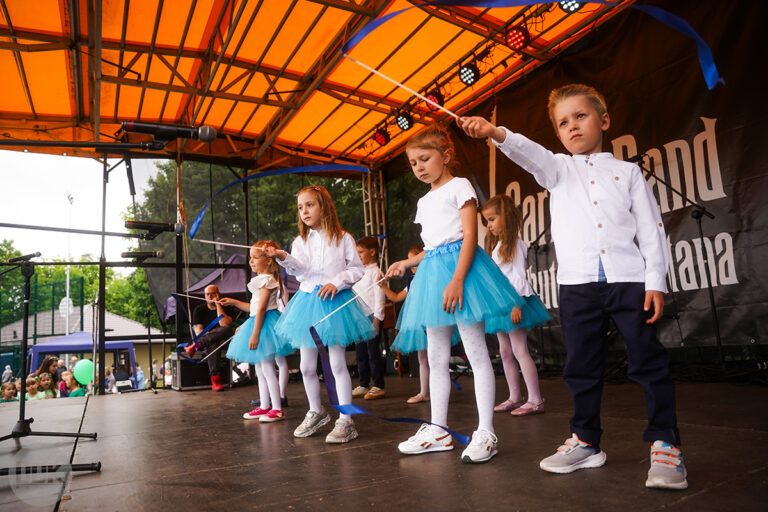 Grupa dzieci stoi na scenie; machają kolorowymi wstążkami.