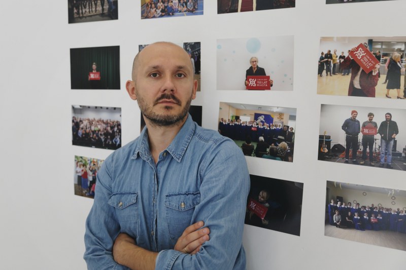 Portret mężczyzny, który zerka w stronę obiektywu, mężczyzna stoi przed ścianą, na której powieszono kilkanaście jego zdjęć.